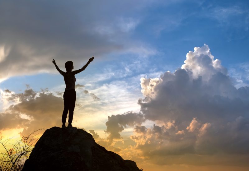 Silhouette of a woman hiker standing alone on big stone at sunset in mountains. Female tourist raising her hands up on high rock in evening nature. Tourism, traveling and healthy lifestyle concept. Silhouette of a woman hiker standing alone on big stone at sunset in mountains. Female tourist raising her hands up on high rock in evening nature. Tourism, traveling and healthy lifestyle concept.